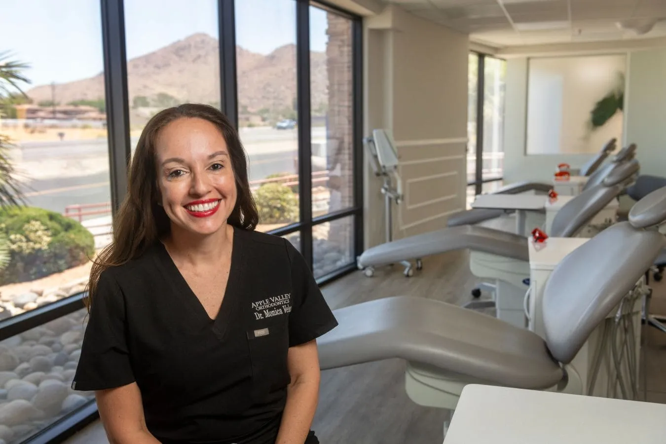 Dr. Monica Velez smiling in a modern, welcoming orthodontic office with treatment chairs and mountain views, representing Apple Valley Orthodontics' patient-focused care and advanced technology.
