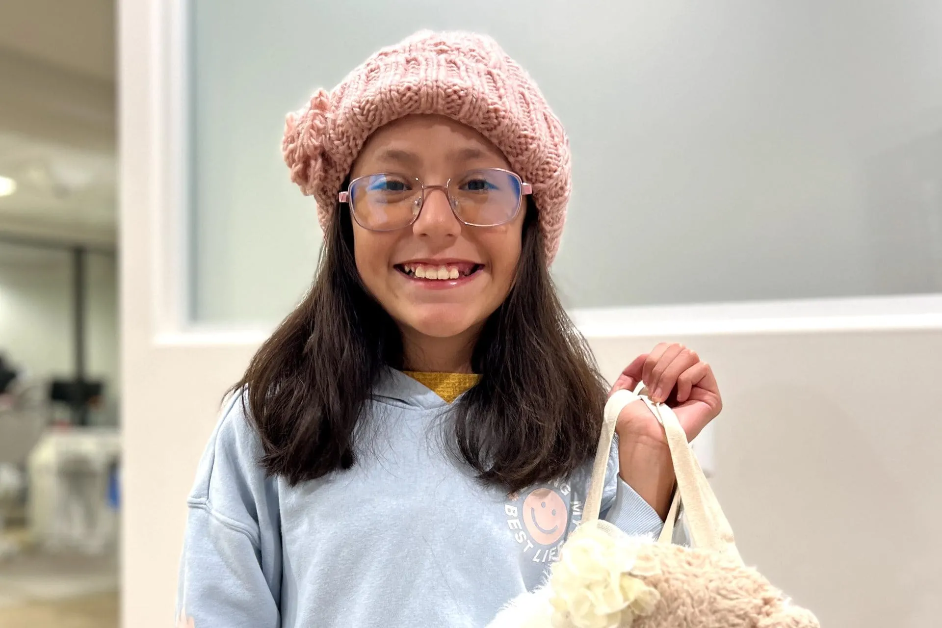 Smiling young girl wearing a pink knit hat and glasses, holding a plush bag, representing the positive effects of early orthodontic treatment for children at Apple Valley Orthodontics.